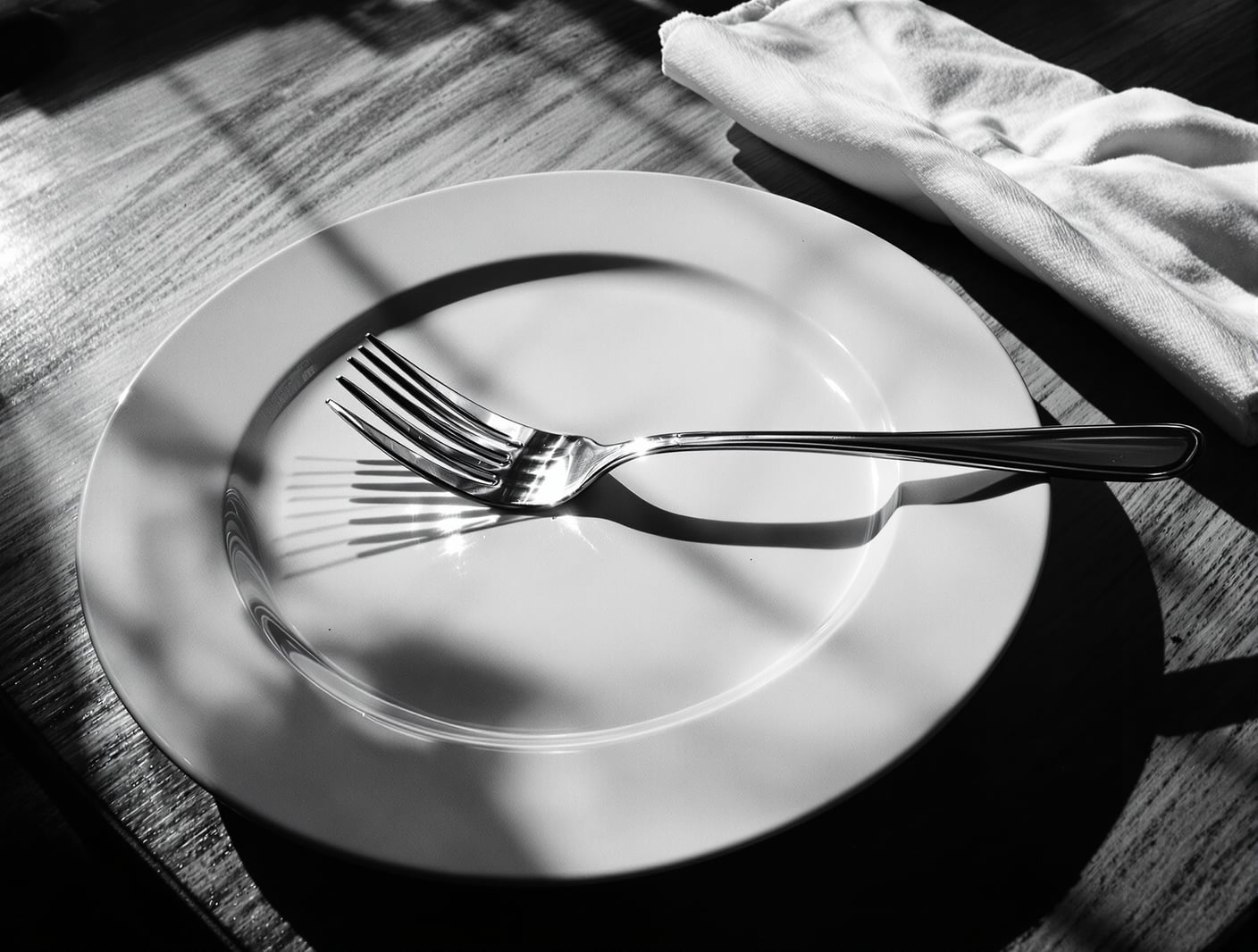 Black-and-white still life shot from the side of a plain ceramic dinner plate with a fork resting across it and a folded