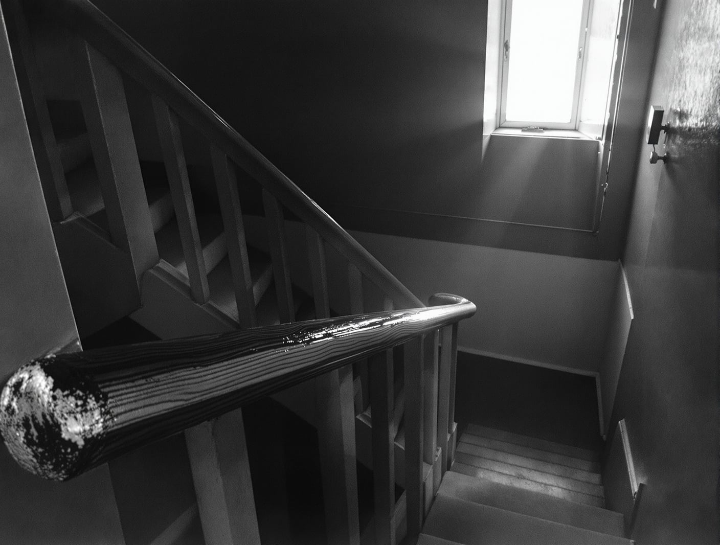 Black-and-white photograph of an empty apartment building stairwell with a worn wooden handrail, hard morning light fall